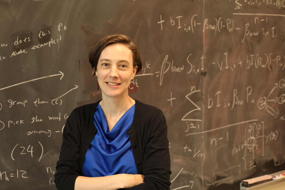 Portrait of Allison with short brown hear, wearing a blue top and black cardigan, standing in front of a chalk board